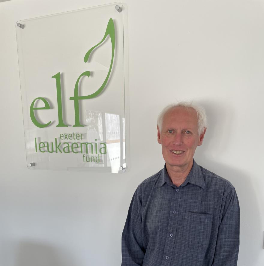 A man smiling stood next to a sign that has the Exeter Leukaemia Fund logo on it